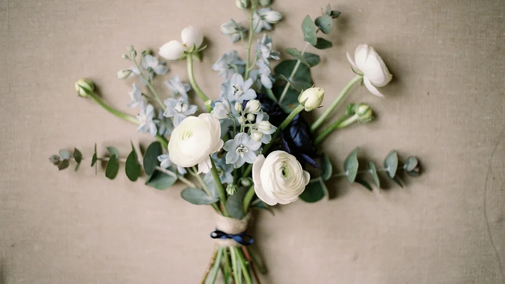 A floral arrangement with white ranunculus, pale blue delphinium, and eucalyptus on a beige linen surface.